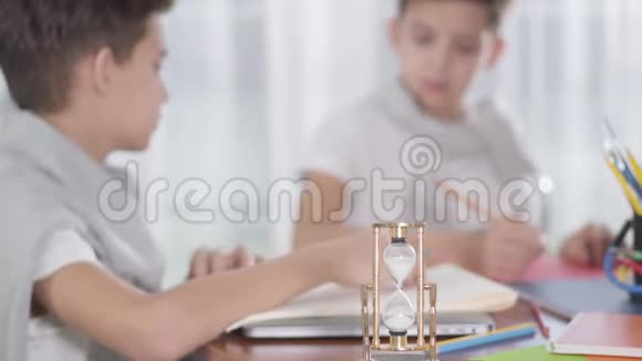 当两个模糊的小学生在背景下画画或写作时旋转沙漏的特写停留在桌子上双胞胎视频的预览图