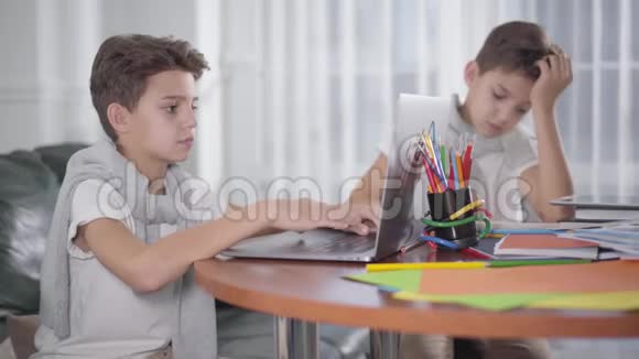 年轻白种人男孩使用笔记本电脑作为他的孪生兄弟在后台写作的肖像兄弟姐妹一起学习视频的预览图