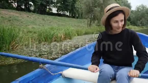 年轻的快乐女孩戴着复古的帽子漂浮在蓝色的木船上湖面上挂着睡莲害羞的女孩在城市公园的湖面上划桨SS视频的预览图