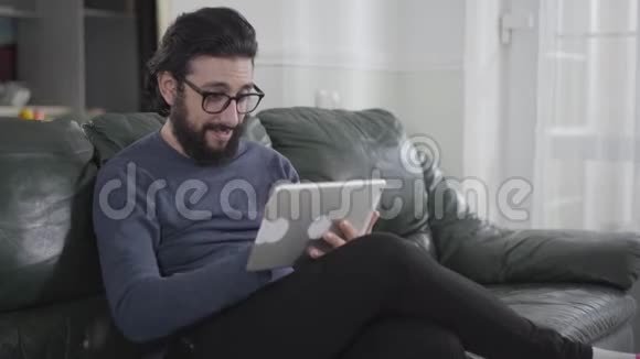 年轻的白种人黑发和胡须戴着眼镜坐在沙发上用平板电脑聪明的家伙视频的预览图