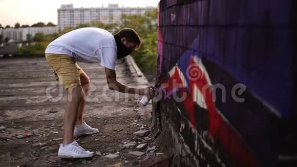涂鸦艺术家画与气溶胶男人穿着巴拉克拉瓦和白色T恤带喷雾瓶年轻的城市画家视频的预览图