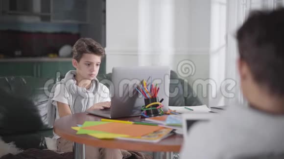 可爱的白种人男孩坐在桌子上拿着笔记本电脑的肖像在家在线学习的学童兄弟模糊或视频的预览图