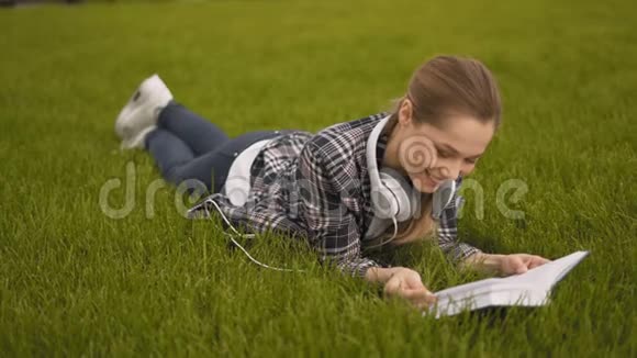格子衬衫大耳机学生女孩躺在公园的草地上看书4K视频的预览图