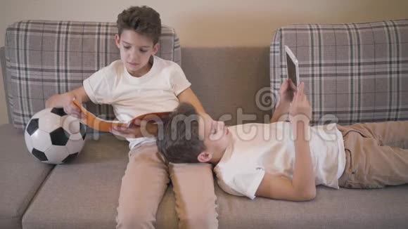 白人男孩坐在沙发上拿着笔记本和球他的孪生兄弟跪着用平板电脑兄弟姐妹视频的预览图