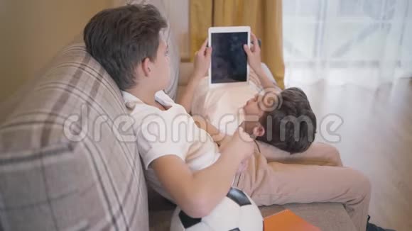 侧面看着黑发白种人男孩与他的孪生兄弟交谈坐在家里的沙发上小兄弟在休息视频的预览图