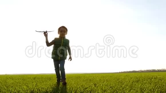 一个快乐快乐的男孩跑过一片绿色的草地手里拿着一架飞机日落背景下快乐的孩子视频的预览图
