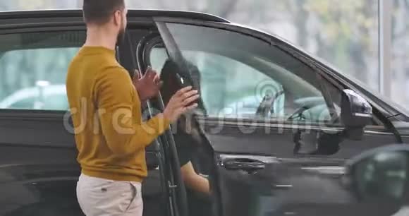 年轻的白种人穿着芥末毛衣打开车门和坐在汽车沙龙里的漂亮女孩交谈夫妇视频的预览图