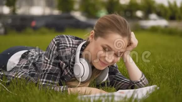 一个女孩看着镜头微笑戴着大耳机一个学生女孩躺在公园的草地上视频的预览图