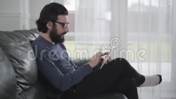 一位自信的高加索商戴眼镜在平板电脑上打字坐在家里年轻人在网上工作视频的预览图
