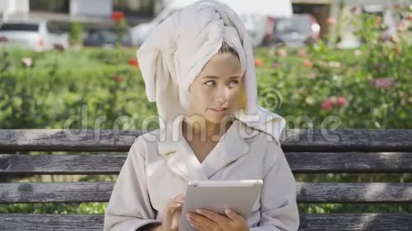 穿着浴袍头上戴着毛巾坐在公园里的长凳上检查的可爱微笑的年轻女子的画像视频的预览图