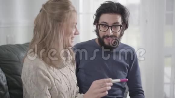 戴眼镜的成年白种人男子看着肯定的怀孕测试微笑着拥抱他可爱的妻子或视频的预览图