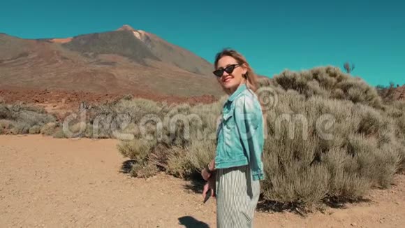 在沙漠火山背景下穿着牛仔夹克条纹裤子和太阳镜的迷人快乐微笑的女人视频的预览图