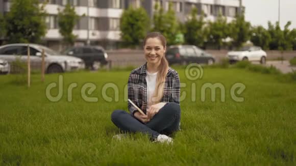 竖起大拇指远射20岁的女孩穿着格子衬衫坐在草地上真诚地微笑着看着摄像机4K视频的预览图