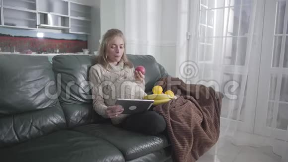 怀孕快乐女孩在平板电脑上看视频和吃苹果的肖像年轻的高加索女人躺在沙发上视频的预览图