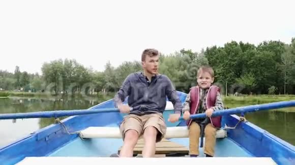 年轻快乐的父亲和他的儿子在绿色公园的木船上快速划船快乐的父亲和小孩一起在湖上度过时光视频的预览图