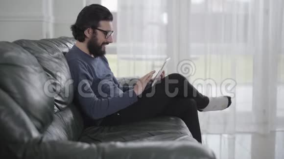 戴眼镜的白种人的侧视图使用平板电脑和微笑年轻人坐在舒适的沙发上视频的预览图
