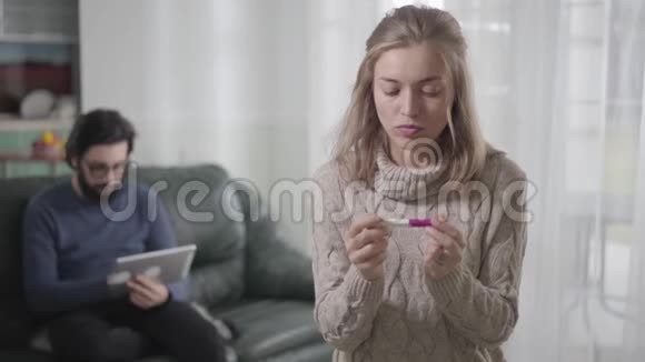 年轻的白种人金发女子持肯定怀孕测试的肖像回顾她的丈夫思考视频的预览图