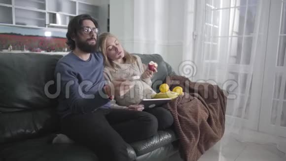 年轻的高加索夫妇坐在家里的沙发上看电视戴眼镜的留胡子的人视频的预览图