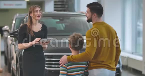 成人白人男子选择汽车与儿子在汽车经销商女汽车经销商持有平板电脑视频的预览图