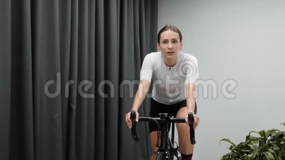 可爱的女人在室内骑自行车穿着白色球衣和粉红色袜子的智能教练在家锻炼心脏视频的预览图