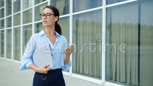 自信的商务女士在玻璃墙办公楼附近使用智能手机视频的预览图