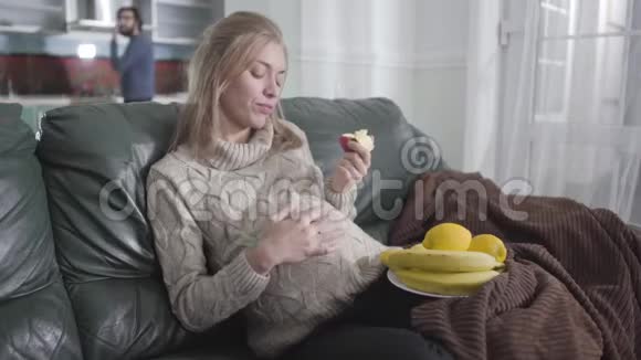 年轻的大胡子男子站在厨房的背景他怀孕的妻子坐在沙发上吃苹果和视频的预览图