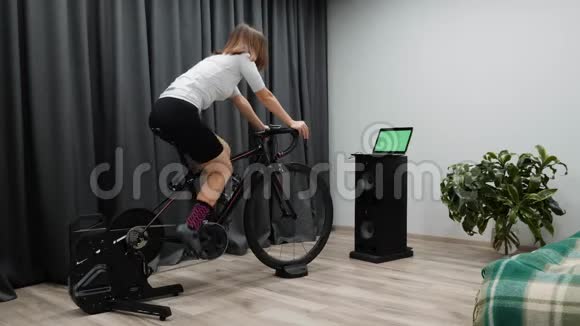 室内自行车概念骑自行车的年轻女性骑在智能教练机上看着电脑屏幕视频的预览图