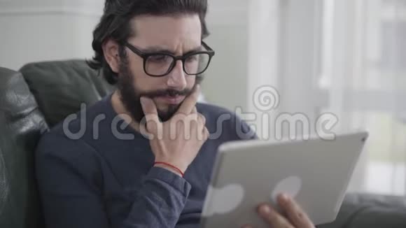 戴着眼镜的自信白种人的特写脸用平板电脑揉着黑色卷曲的胡须视频的预览图