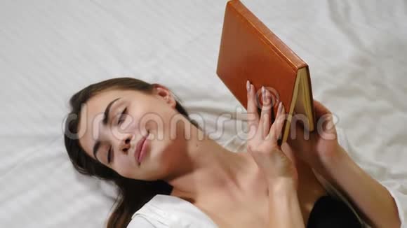 年轻漂亮的女人躺在床上手里拿着一个紧闭的笔记本穿白色衣服的轻松迷人的女士视频的预览图