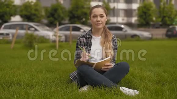 很长一段时间学生女孩坐在草地上在笔记本上做笔记这个女孩解决了一项非常重要的任务4K视频的预览图