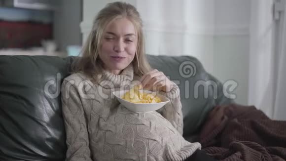 年轻孕妇看相机和吃薯片的特写肖像金发白种人女孩坐在沙发上视频的预览图