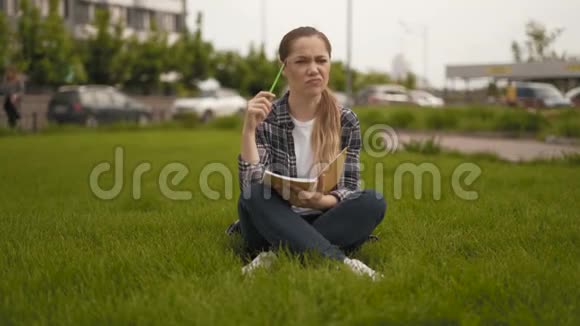 我有个主意远射20岁的女孩穿着格子衬衫坐在草地上在笔记本上记笔记视频的预览图