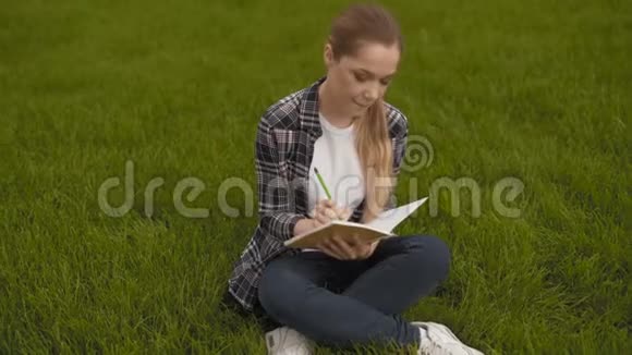 穿格子衬衫的少女坐在草地上在笔记本上做笔记4K视频的预览图