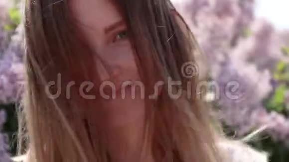 在一个阳光明媚的日子里年轻的金发女孩在紫丁香的背景上看着相机视频的预览图