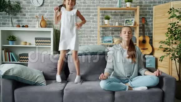 快乐的小女孩在沙发上跳而妈妈则在家里的莲花位置放松视频的预览图