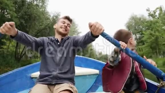 快乐的父子在绿色公园的木船上划船年轻快乐的父亲和他可爱的小儿子坐船在湖上航行视频的预览图