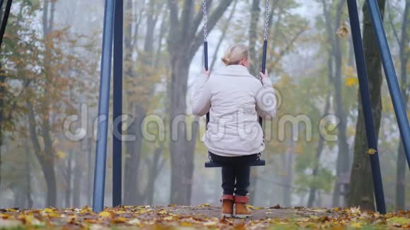 在有浓雾的公园里一个女人坐在秋千上的后视镜孤独概念视频的预览图