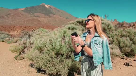 女人穿着牛仔夹克和太阳镜用手机拍自然照片火山背景下的沙漠视频的预览图