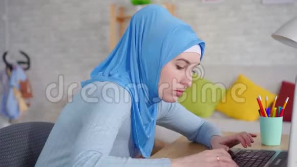 年轻的穆斯林妇女戴着传统的头巾坐在笔记本电脑前感到脖子上的疼痛视频的预览图