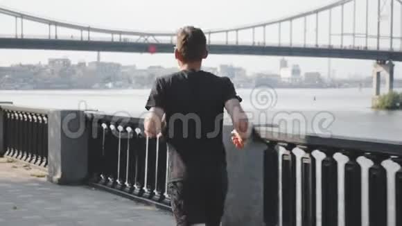 活泼苗条的男人穿着黑色t恤和运动短裤沿着河跑年轻的职业运动员在城市跑步健康强视频的预览图