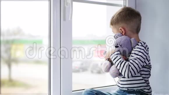 一个小男孩坐在窗边抱着一个毛绒玩具孩子看着窗外很伤心视频的预览图