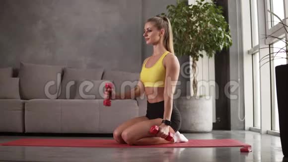 年轻的金发女郎坐在地板上的垫子上锻炼在宽敞的客厅里举起红色的哑铃视频的预览图