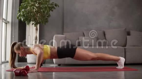 穿运动服装的健康年轻女性黄色上衣和黑色短裤在灰色沙发前的健身垫上做俯卧撑视频的预览图