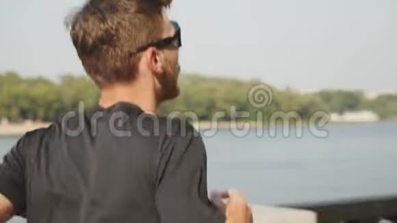 性感苗条的白种人在河边跑穿着黑色t恤和运动眼镜的年轻运动员早上在城里跑步氯视频的预览图