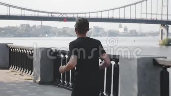 年轻活泼的白种人穿着黑色T恤和短裤沿着城市码头奔跑运动服运动员在城市跑步健康青年视频的预览图