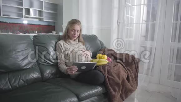 快乐怀孕的白种人女人在平板电脑上看视频和抚摸她的腹部的肖像年轻女孩躺在视频的预览图