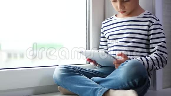 可爱的男孩坐在白色的窗户上玩平板电脑视频的预览图