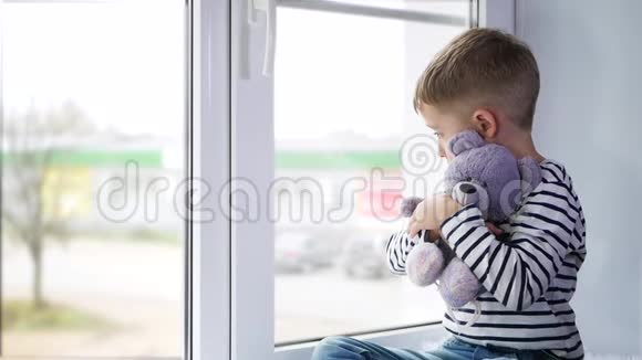 一个悲伤的小男孩抱着一只常春藤泰迪熊在阴天坐着看着窗外视频的预览图