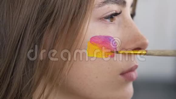 一个年轻漂亮女孩的特写肖像艺术家用小软笔在女孩脸上画多色图标视频的预览图