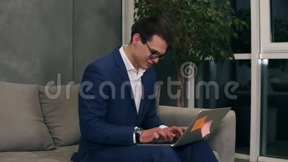 白种人商人穿着正式的蓝色西装戴着时髦的眼镜坐在沙发上跪在笔记本电脑上打字视频的预览图